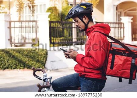 Courier in red uniform with a delivery box on back riding a bicycle and looking on the cellphone to check the address to deliver food to the customer. Courier on a bicycle delivering food in the city.
