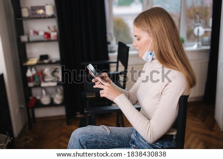 Female customer in beauty salon sitting on hight chair in medical mask on chin and use phone. Beautiful young blond hair woman wiating for brow master Zdjęcia stock © 