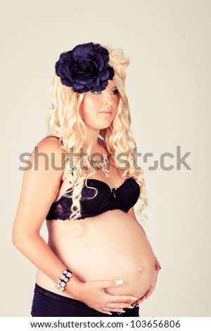 Beautiful pregnant woman with long curly blonde hair and a very large protruding belly standing in black lingerie holding her stomach