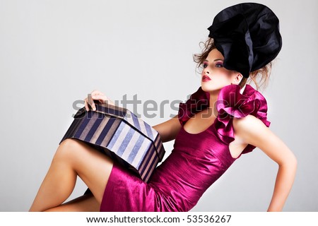 A young woman dressed in avant garde attire and holding a hat box. She is wearing a hat and has cosmetic artwork on her right temple. Horizontal shot.