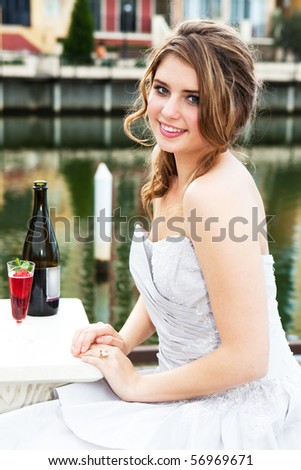 An attractive young woman is smiling and wearing formal attire while sitting at a dockside table with an alcoholic beverage. Vertical shot.