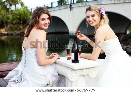 Two attractive young women are wearing formal attire and sitting on a dock sharing a drink. Horizontal shot.