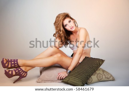 Attractive young woman wearing lingerie and high heels sits on a stack of pillows while looking into the camera. Horizontal shot.