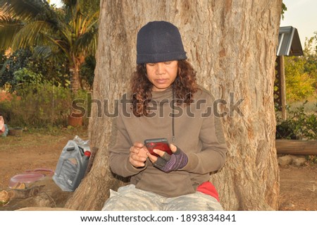 Real farmer thai woman , Asian aged 50 years chatting  the smartphone in hand dressed like a soldier, seating relax on the log and big tree behind. Stock fotó © 