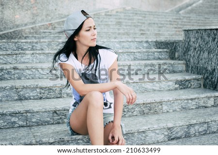 Young teenage girl with headphones,black long hair and jeans shorts sitting on steps and attentively looking right.