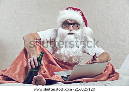 Bearded man in Santa hat drinking beer in the bed with laptop