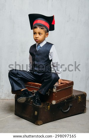 small black boy in suit and graduation hat sits on the old suitcases and look left.  instagram toned