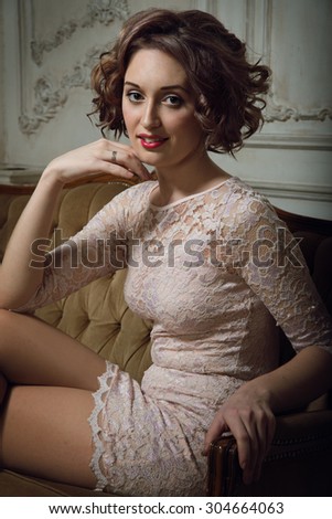 Curly brunette in pink dress sit at the sofa.