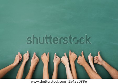 Close up view of the hands of a group of people giving a thumbs up gesture of approval an success with their hands raised against a blank green chalkboard with copyspace