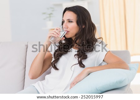 Pretty brunette drinking water on couch at home in the living room