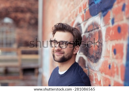 Portrait of Smiling Young Man with Facial Hair Wearing Eyeglasses and Leaning Against Brick Wall Painted with Graffiti