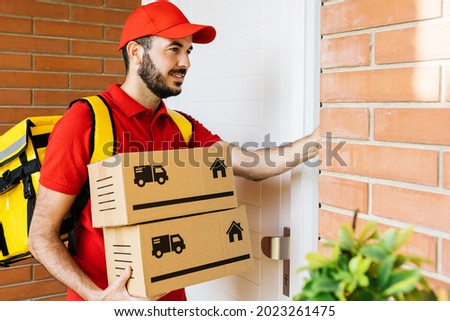Happy delivery man in red uniform with backpack ringing the house doorbell while holding cardboard box - Hispanic courier rider delivering a package - Small business and delivery service concept