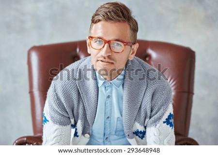 Portrait of a young intelligent man stares into the camera, good view, small unshaven, charismatic, wearing glasses rimmed with brown, blue shirt, gray sweater, sitting on a brown leather chair