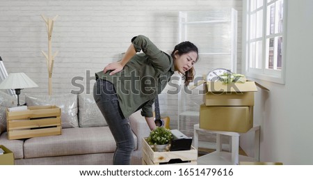 beautiful young single woman is moving in new apartment. girl having pain in back while packing standing among cardboard boxes. female injured of waist while carrying heavy wood container with stuff 商業照片 © 