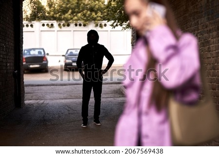 Criminal stalking a woman alone in dark street alley. Female is talking on phone. Unrecognizable man is hiding in the shadows. Woman Doesn't Suspecting That She Is Being Followed By Criminal Bandit Stockfoto © 