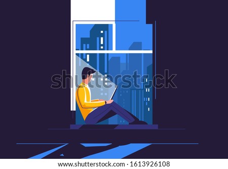 A young man is sitting on the windowsill with a laptop, in the dark against the background of the night city outside the window