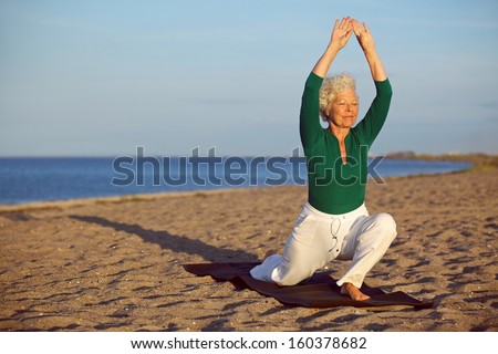 Senior woman practicing yoga poses on the sandy beach. Elder caucasian woman stretching legs and arms on the seashore. Healthy lifestyle and fitness concept