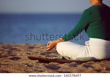 Cropped image of senior woman in meditation on sandy beach. Elderly woman sitting on the beach in lotus pose doing relaxation exercise . Mature woman doing yoga
