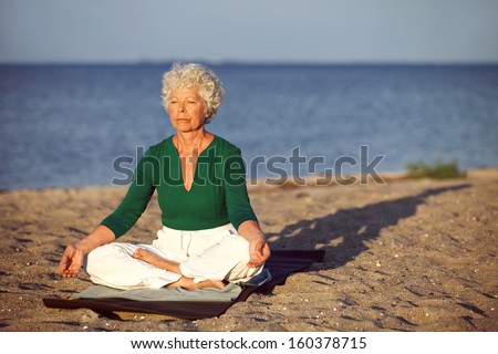Elderly woman sitting on exercise mat doing meditation in lotus pose on the beach. Old woman doing relaxation exercise on the seashore. Meditation, yoga and relaxation concept