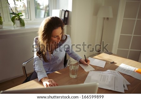 Young businesswoman sitting at her desk writing notes from a laptop at home. Beautiful caucasian female working from home office.