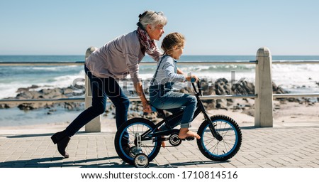 Side view of a girl riding a bicycle while her grandmother runs along holding the kid. Senior woman teaching a small girl to ride a bicycle on the road alongside the beach.