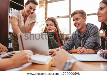 Female teacher working with college students in library. Group of students studying in school library with a teacher helping them.