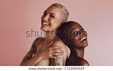 Two women with buzz cut hairstyle standing back to back and smiling at camera. Female models with beautiful skin smiling together over beige background.