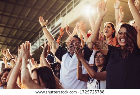 Crowd of sports fans cheering during a match in stadium. Excited people standing with their arms raised, clapping and yelling to encourage their team.