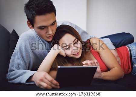 Lovely teenage couple lying together on sofa using tablet PC. Mixed race couple relaxing on couch surfing internet on digital tablet computer at home.