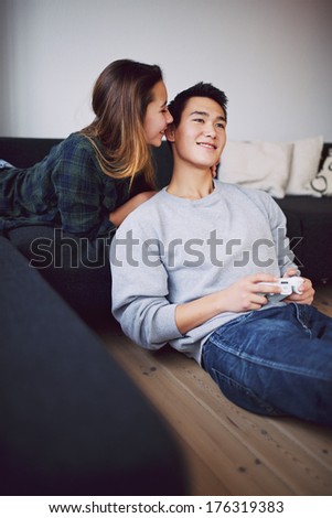 Handsome young man playing video game while his girlfriend whispering something in his ears. Teenage asian couple relaxing in living room at home.