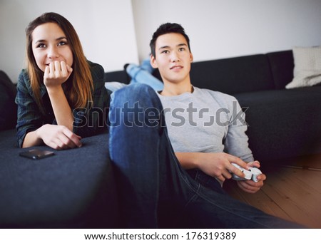 Beautiful young asian woman lying on sofa while young man sitting on floor playing video game at home. Mixed race teenage couple relaxing indoors.