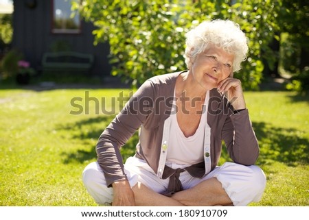 Senior woman sitting outdoors on grass looking away and thinking. Elder woman relaxing in backyard garden day dreaming