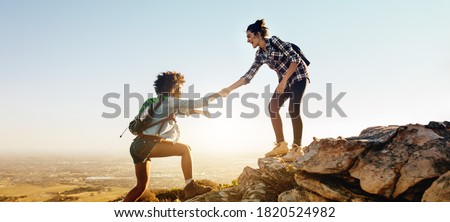 Woman helping her friend to climb the cliff and reach the top of mountain. Friends helping each other during hiking a mountain.
