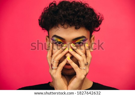 Gender fluid male wearing rainbow colored eye shadow and smiley face on fingernail. Transgender male with funky makeup on red background.