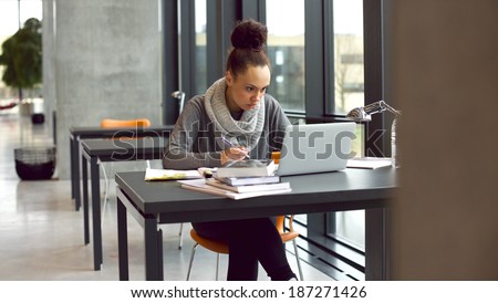Young afro american woman sitting at table with books and laptop for finding information. Young student taking notes from laptop and books for her study in library.