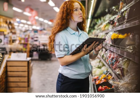 Woman using a digital tablet while doing inventory check in a grocery store. Female trainee employee working in supermarket.