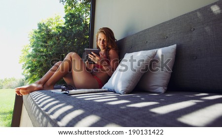 Beautiful and happy young lady in bikini sitting on a couch using digital tablet. Female model working on tablet computer outdoors.