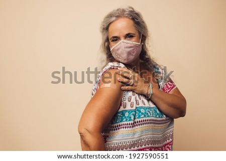 Portrait of a senior woman with protective face mask showing her arm with bandage after getting vaccine. Mature woman sitting against brown background after receiving corona virus vaccination.