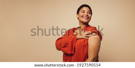 Woman showing her arm after receiving vaccine shot on brown background. Female with bandage on her arm looking away and smiling.