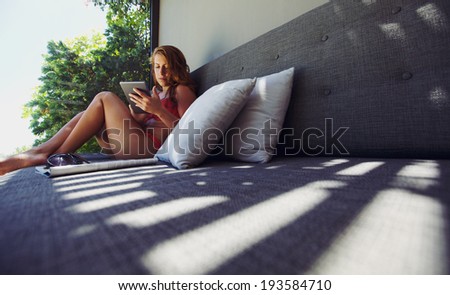 Attractive young woman in bikini sitting on a couch using digital tablet. Female model surfing the net with tablet computer outdoors.
