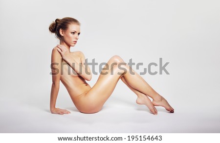Sensual nude woman sitting on floor covering her breast with her hand and looking away at copyspace. Pretty young nude woman sitting on grey background.