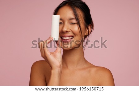 Pretty woman model holding a cosmetic bottle and smiling. Playful Asian female with new skincare product.