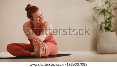 Smiling woman sitting on fitness mat and stretching leg. Fit female doing warmup workout at fitness studio.