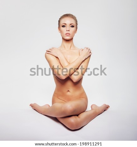 Young naked girl in an artistic sitting yoga pose. Nude caucasian female sitting on floor with her arms and legs crossed on grey background.
