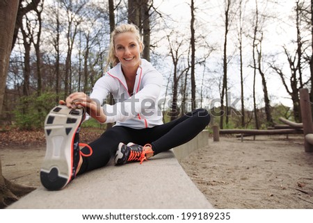Happy and fit woman stretching her legs before training session in nature. Beautiful caucasian athlete exercising at the park smiling at camera.