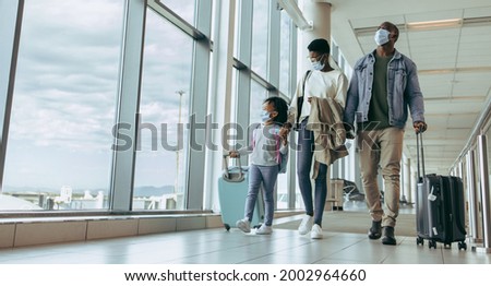 African family with luggage walking by window at airport terminal and watching airplanes through glass window. Family walking through airport passageway.