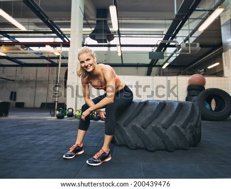 Woman sitting on tire and smiling at camera at gym. Crossfit female athlete taking rest after working out.