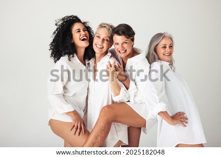 Photo of Female models of different ages celebrating their natural bodies in a studio. Four confident and happy women smiling cheerfully while wearing white shirts against a white background.