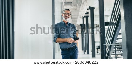 Smiling mature businessman holding a smartphone in an office. Businessman looking at the camera while standing alone in a modern workplace. Experienced businessman communicating with his clients.