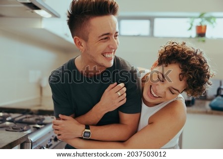Young queer couple laughing together indoors. Happy young queer couple having fun together while standing in their kitchen. Romantic young LGBTQ+ couple bonding fondly at home.